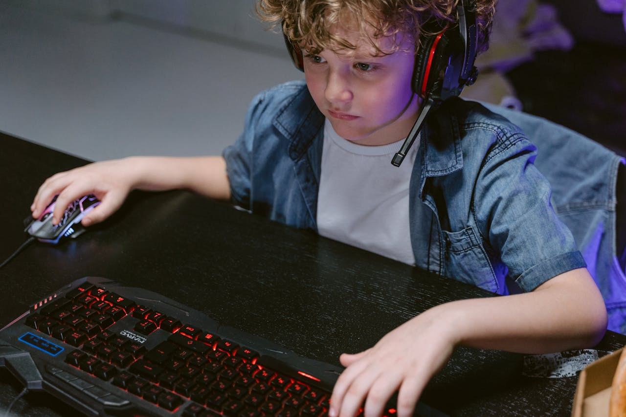 A young child using a keyboard and mouse with headphones, playing a computer game indoors.