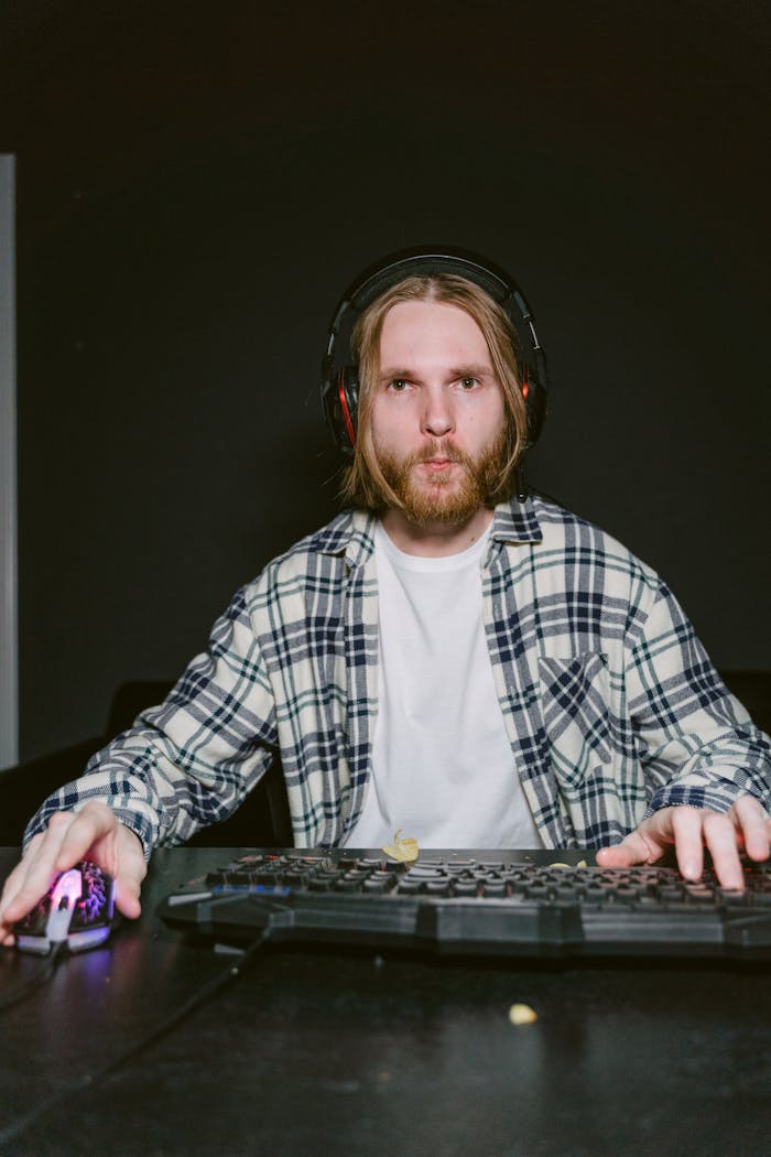 Man in plaid shirt wearing headphones, deeply focused on gaming at a computer.