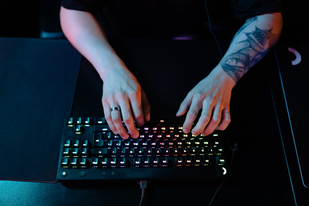 Close-up of hands typing on a RGB gaming keyboard with a tattooed arm and ring visible.